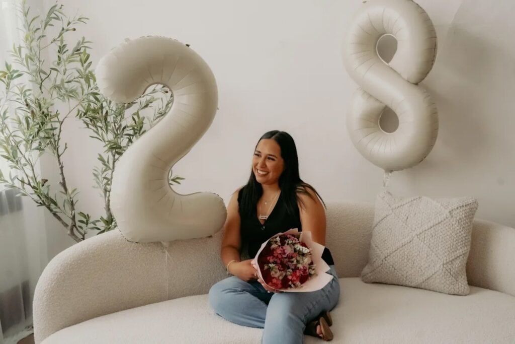 Woman sits on couch with flowers surrounded by birthday balloons for her 28th birthday