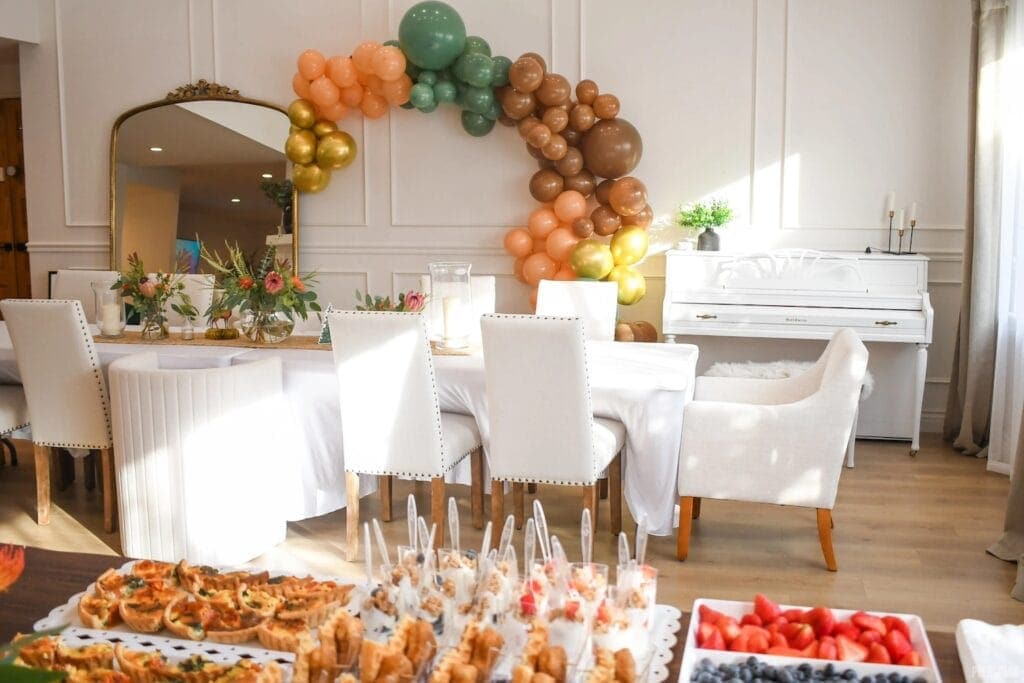 White chairs surround long table set with white tablecloth and flowers with a balloon arch in the background and snacks in the foreground.