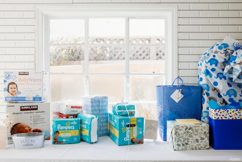Diapers, baby wipes and gifts wrapped in blue paper sit on a gift table by a window at a baby shower