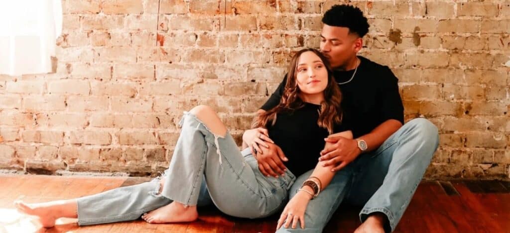 Couple sitting on floor in matching outfits leaning against brick wall.