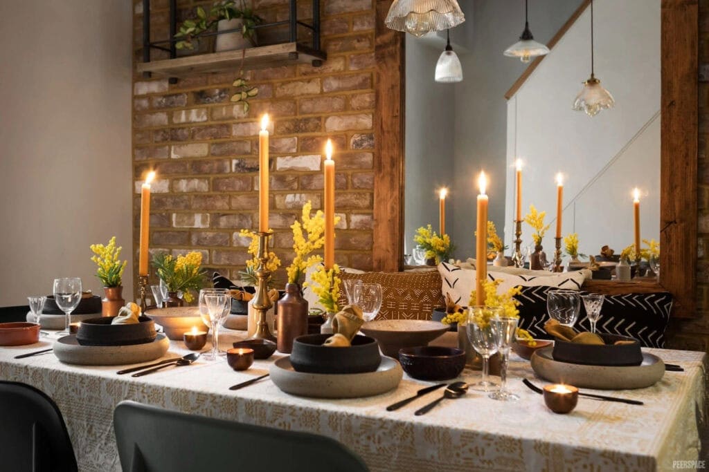 A small dining table is set in earth tones by a brick wall with a large mirror reflecting the tablescape