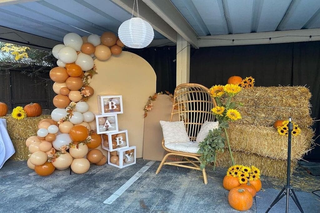 A wooden chair sits next to hay bales and orange-hued balloons at a fall-themed baby event
