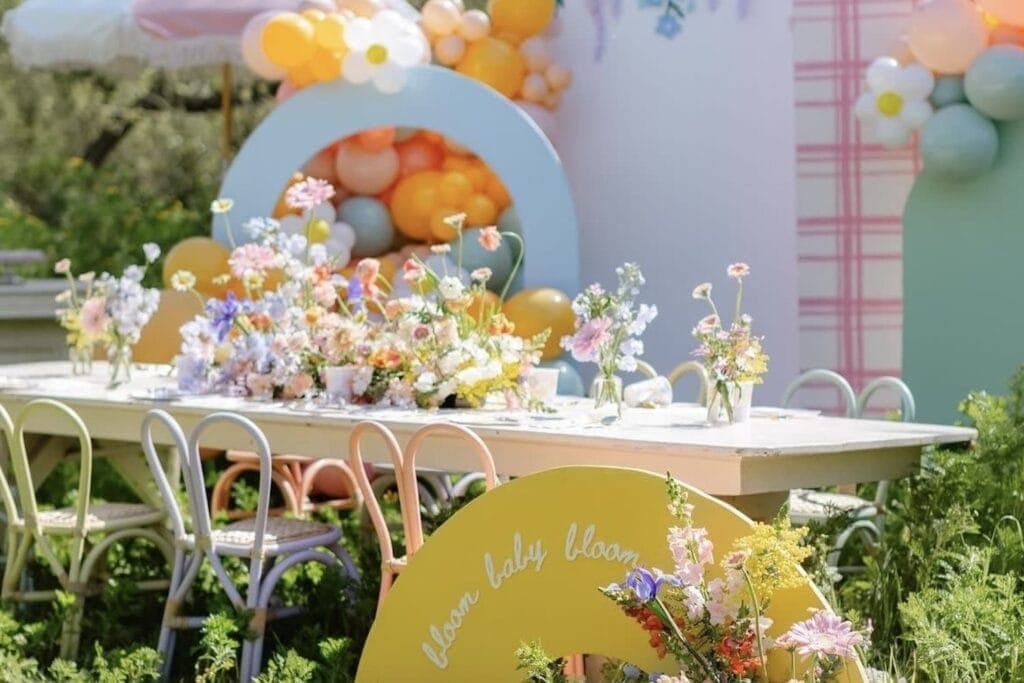 Spring flowers sit on a table set up outside with pastel chairs and a "Baby in Bloom" sign in the forefront