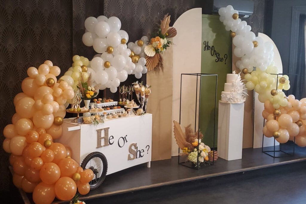 A gender neutral display of balloons surround a cake and dessert table at a gender reveal party