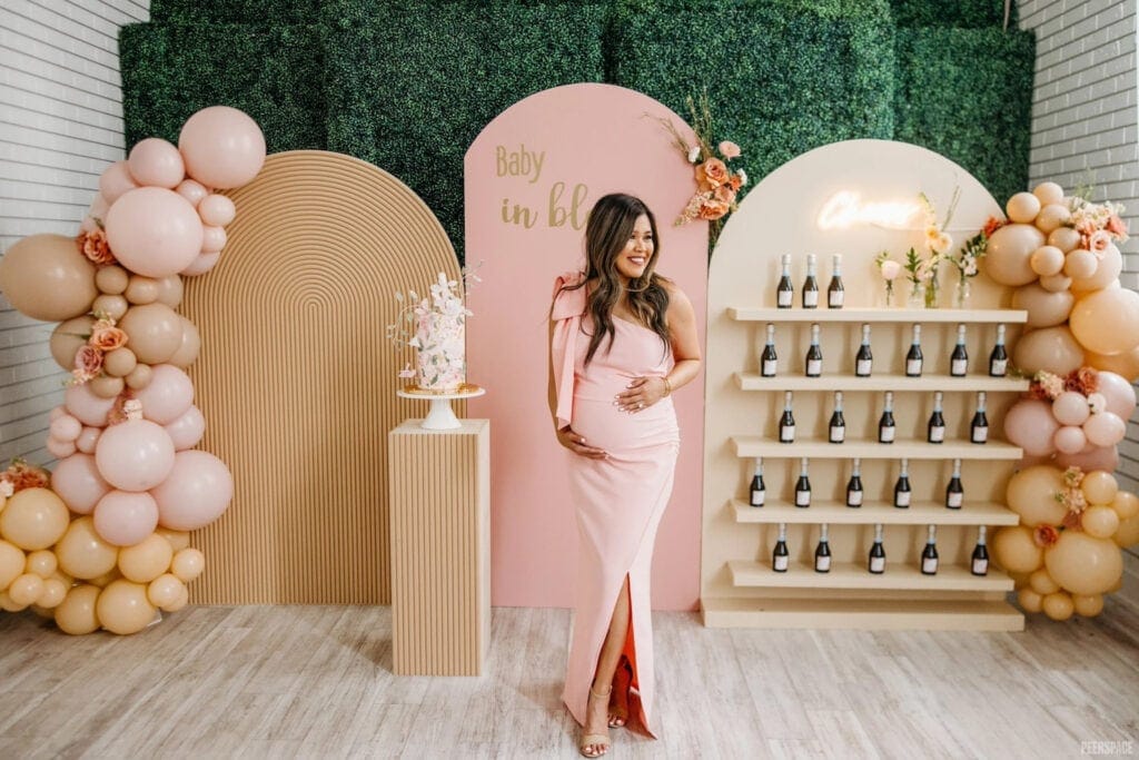 A mother to be in a pink dress poses in front of baby party decor and a wall of champagne