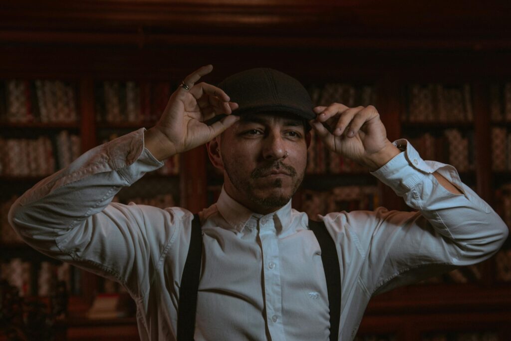 A man in suspenders and a white collared shirt adjusts his newsboy cap