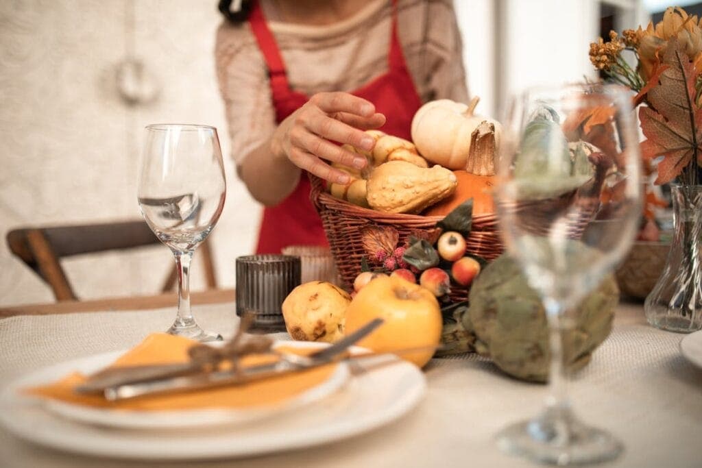 Person decorating a fall-themed Friendsgiving table with tablecloth, basket of pumpkins, foliage, and dishware