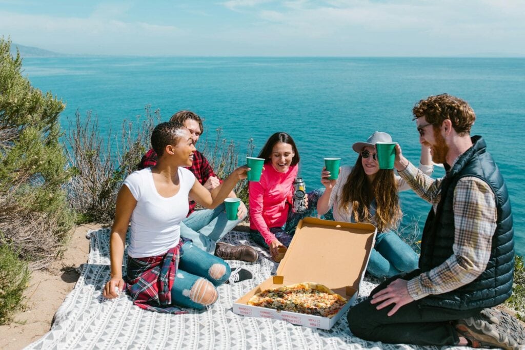 Five friends having a picnic birthday party with pizza near the coast